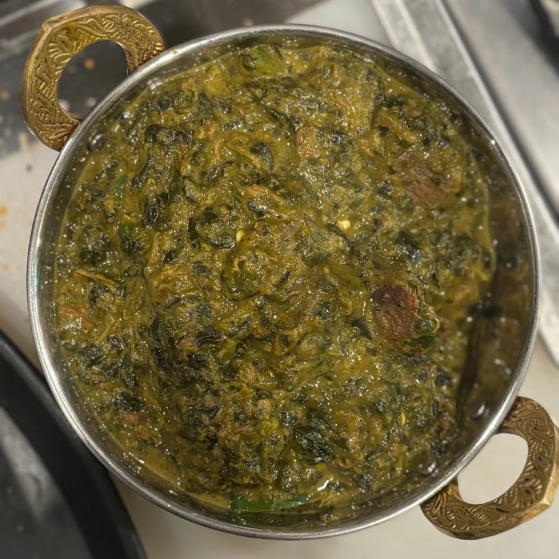 Overhead view of a metal bowl with ornate brass handles filled with green saag.